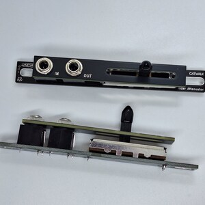 May include: A black electronic audio device with a slider and two 1/4 inch jacks labelled "IN" and "OUT". The device is labelled "CATWALK Slider Attenuator".
