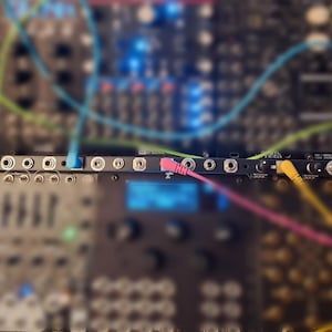 May include: Close-up of a black modular synthesizer with a row of jacks and patch cables in various colours, including red, blue, green, yellow, and pink.