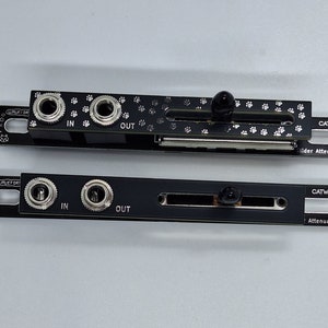 Puede incluir: Dos placas de circuito impresas negras con conectores plateados etiquetados como "IN" y "OUT". La placa superior tiene un control deslizante con una perilla negra y el texto "CATWALK Slider Attenuator". La placa inferior tiene el texto "CATWALK Attenuator".