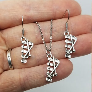 May include: A silver necklace and earrings set featuring a xylophone charm. The charm has a silver xylophone with two silver mallets crossed over the xylophone.