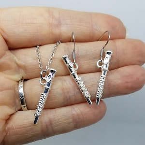 May include: Silver flute necklace and earrings set. The necklace features a silver flute pendant on a silver chain. The earrings are silver flute charms on silver hooks.