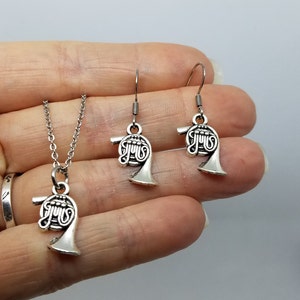 May include: A silver necklace and earrings set featuring a French horn charm. The necklace has a silver chain and the earrings are dangle style.