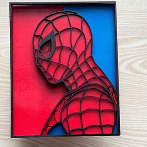 May include: A black framed wall art piece featuring a red and blue background with a black silhouette of Spider-Man's head and shoulders. The silhouette is cut out to reveal the red and blue background.