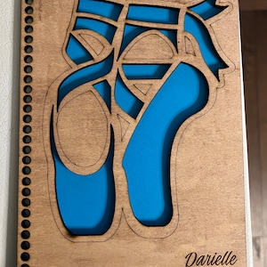 May include: A wooden cutout of a pair of ballet shoes with a blue background. The cutout is on a wooden board with the text "Darrielle" engraved on the bottom.