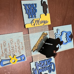 May include: A blue and yellow graduation box with the words "You Did It!" and "Class of 2024" on it. The box is decorated with a graduation cap, a diploma, and a silhouette of a person in a graduation gown. The name "Maya" is written on the box.