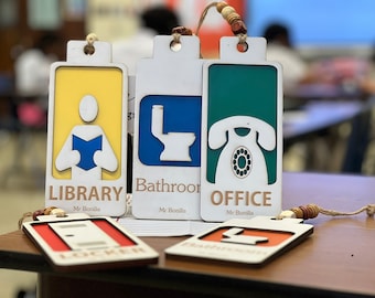 May include: Five wooden signs with different icons and text. The signs are yellow, blue, green, red, and white. The icons are a person reading a book, a toilet, a phone, a door, and a toilet. The text on the signs is "LIBRARY", "Bathroom", "OFFICE", "LOCKER", and "Bathroom".