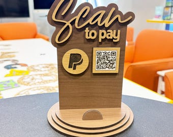 May include: Wooden "Scan to Pay" sign with a PayPal logo and a QR code. The sign is made of layered wood, featuring a dark brown base and lighter wood accents for the text and logo. The sign is designed to stand on a counter.