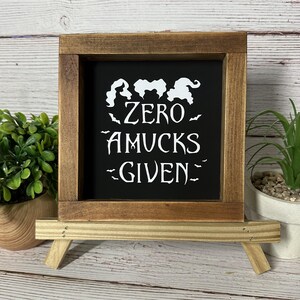 May include: A wooden framed sign with the text "ZERO AMUCKS GIVEN" in white on a black background. The sign is propped up on a small wooden easel. Decorative plants are on either side of the sign.
