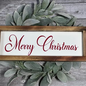 May include: A rectangular wooden sign with the words "Merry Christmas" in red script on a white background. The sign is framed in brown wood and rests on a bed of green and gray leaves, set against a gray wooden background.
