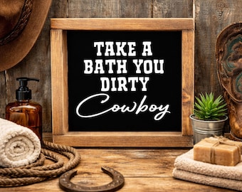 Op de afbeelding: Een houten bord met de tekst "TAKE A BATH YOU DIRTY Cowboy" in witte letters op een zwarte achtergrond. Het bord staat op een houten oppervlak met een cowboyhoed, laarzen en badaccessoires.
