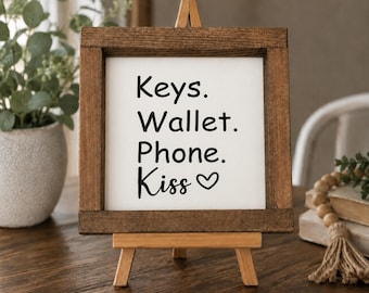 May include: A square wooden sign with a dark brown frame and a white center panel. The text printed in black reads: Keys. Wallet. Phone. Kiss followed by a small heart symbol. The sign is displayed on a small, light-colored wooden easel stand.