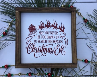 May include: A square wooden sign with a dark brown frame and a white center panel. The sign features dark red text that reads, "May you never be too grown up to search the skies on Christmas Eve," accompanied by a silhouette of Santa in a sleigh.