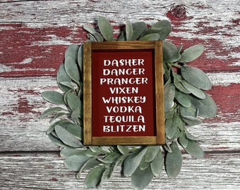 May include: A framed sign with a red background and white text listing "Dasher, Dancer, Prancer, Vixen, Whiskey, Vodka, Tequila, Blitzen." The sign is surrounded by a wreath of green leaves, set against a rustic, red and white wooden background.
