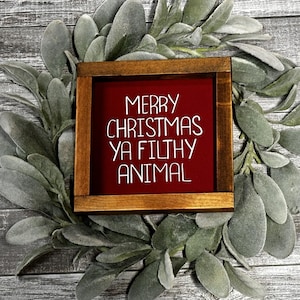 May include: A wooden framed sign with the text "MERRY CHRISTMAS YA FILTHY ANIMAL" in white on a red background. The sign is surrounded by a wreath of green and gray leaves, set against a gray wooden background.