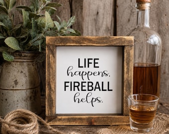 May include: A square wooden sign with a dark brown frame and a white center panel featuring the black text "LIFE happens. FIREBALL helps." The sign is positioned next to a clear glass bottle filled with amber liquid and a small matching shot glass.