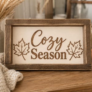 May include: A rectangular wooden sign with a dark brown stained frame and a light-colored center panel. The text "Cozy Season" is engraved in the center, flanked by two detailed maple leaf illustrations on the left and right sides.