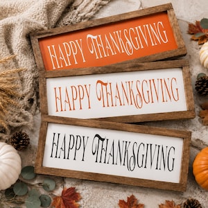May include: Three rectangular wooden signs with dark brown frames, each featuring the text "HAPPY THANKSGIVING" in a decorative font. The top sign has white text on an orange background, the middle has orange text on white, and the bottom has black text on white.