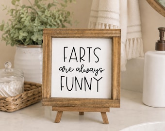May include: A square wooden sign with a natural brown finish and a white center panel. The sign features black text that reads "FARTS are always FUNNY" in a mix of sans-serif and script fonts. The sign is displayed on a small wooden easel stand.