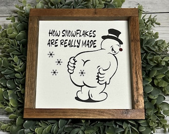 May include: A square wooden sign with a dark brown frame featuring a white center panel. The text reads HOW SNOWFLAKES ARE REALLY MADE above a black and white illustration of a snowman wearing a top hat, bending over to produce snowflakes.