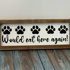 May include: A rectangular wooden sign with a white background and a brown frame. The sign features five black paw prints above the phrase "Would eat here again!" in black script. A small potted plant is to the right.