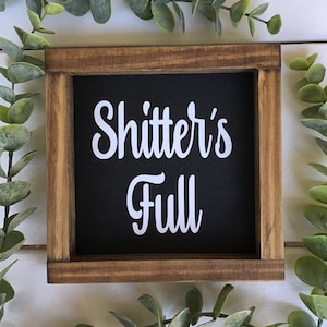 May include: Square wooden sign with a black background and the words "Shitter's Full" in white script. The sign has a brown wooden frame and is surrounded by green foliage.
