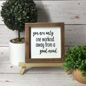 May include: A small wooden sign with the text "you are only one workout away from a good mood" in black lettering. The sign is displayed on a small wooden easel. The sign is surrounded by decorative plants.