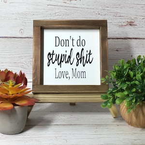 May include: A framed sign with the text "Don't do stupid shit Love, Mom" in black lettering. The sign is in a brown wooden frame and is displayed on a wooden stand. Decorative plants are in the foreground.