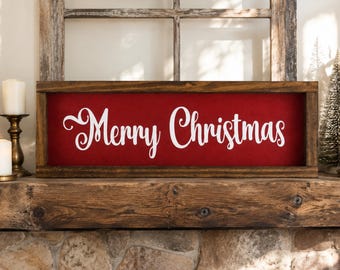 Puede incluir: Un letrero rectangular de madera con un marco marrón oscuro y un fondo rojo intenso. El texto Merry Christmas está escrito en letra cursiva blanca en el centro del letrero. El letrero está colocado horizontalmente sobre una repisa de madera.