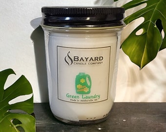 May include: A glass jar candle with a black lid. The label reads "BAYARD CANDLE COMPANY" and features a graphic of a green laundry detergent bottle with the words "Green Laundry". Made in Webberville, MI.