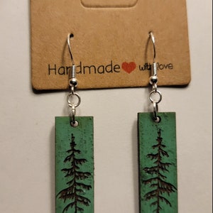 May include: A pair of green wood earrings with a silver hook and a laser-etched pine tree design. The earrings are on a brown card with the text "Handmade with love".