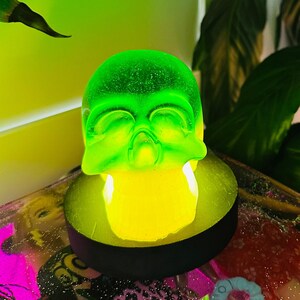 May include: A green glass skull shaped night light with a yellow glow. The light is sitting on a black base and is on a table with a pink and white background.