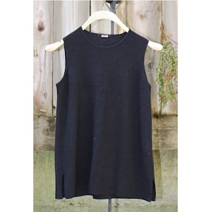 May include: A sleeveless black top hangs on a wooden hanger. The top has a round neckline and side slits. The fabric appears to be a soft knit material. The background is a weathered wooden fence.