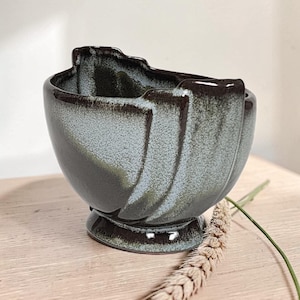 May include: A ceramic bowl with a pedestal base, featuring a glossy glaze in shades of dark brown and muted sage green. The exterior surface has a textured, speckled appearance with vertical molded ridges that extend toward the uneven rim.