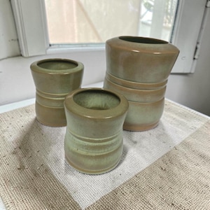 May include: A set of three ceramic vessels with a smooth, matte finish in muted shades of sage green and tan. Each vessel features a cylindrical shape with a slightly flared rim and a subtle horizontal indentation around the center of the body.
