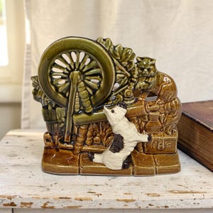 May include: A glazed ceramic figurine featuring a spinning wheel in olive green, a brown cat perched on a wooden frame, and a white dog with black spots standing in the foreground. The base is brown with the letters R H S embossed on a small box.
