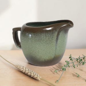 May include: A ceramic creamer pitcher featuring a smooth, dark brown rim that transitions into a speckled, light green body. The pitcher has a curved handle on the left side and a small pouring spout on the right, with a glossy finish throughout.