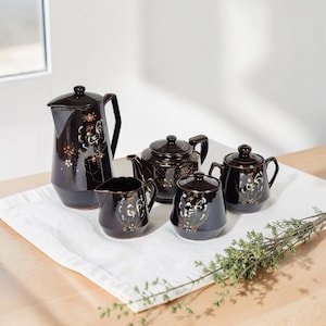 May include: A dark brown ceramic tea set consisting of a tall pitcher, a teapot, a creamer, and a lidded sugar bowl. Each piece features a hand-painted rooster motif in white, orange, and gold tones with delicate floral accents and gold trim.