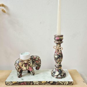 May include: A floral patterned elephant candle holder and a tall candlestick. The candlestick holds a beige candle and is tied with twine. The elephant holds a small white candle. Both are on a decorative wooden base, with a floral pattern.