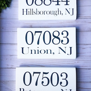 May include: Three white wooden signs with black painted numbers and text. The top sign reads "08844 Hillsborough, NJ". The middle sign reads "07083 Union, NJ". The bottom sign reads "07503 Paterson, NJ".