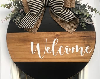 May include: A round wooden door hanger with a black and brown striped design. The word "Welcome" is written in white on the wooden portion. The hanger is decorated with a burlap bow and greenery.