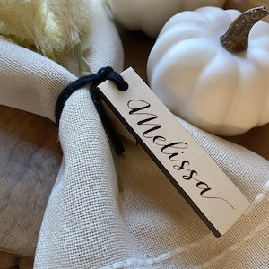 May include: A wooden place card with the name "Melissa" in elegant script. The card is attached to a cream-colored napkin with a black cord. White pumpkins are in the background, suggesting a fall or Thanksgiving theme.