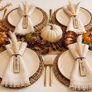 May include: A table setting featuring four cream-colored plates on woven round placemats. Each plate has a fringed napkin tied with a dark ring, a dried grass sprig, and a wooden tag inscribed with the words family, blessed, thankful, or gather.