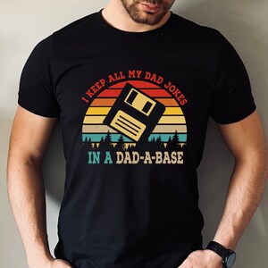 May include: A black t-shirt with a retro sunset graphic and a floppy disk. The text on the shirt reads "I keep all my dad jokes in a dad-a-base."