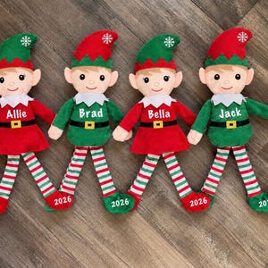 May include: Four plush elf dolls with red and green outfits, white collars, and striped legs. Each elf features a name embroidered on the chest: Allie, Brad, Bella, and Jack. The year 2026 is printed on one foot of each doll, and a snowflake is on each hat.