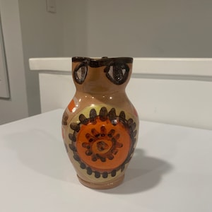 May include: A brown ceramic pitcher with a geometric design in orange, yellow, and black. The pitcher has a rounded body and a wide mouth.