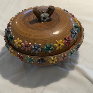 May include: A brown ceramic trinket dish with a lid. The lid has a wooden handle and is decorated with a ring of colorful flowers. The dish itself is also adorned with a row of flowers in various colors.
