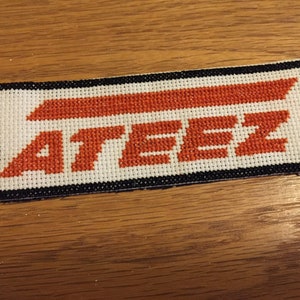 Può includere: Un toppa di punto croce bianco e arancione con la parola "ATEEZ" in lettere arancioni.