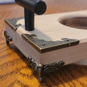 Pu&ograve; includere: Una scatola di legno con una maniglia nera e angoli e piedini in ottone decorati. La scatola ha un ritaglio rotondo nella parte superiore.