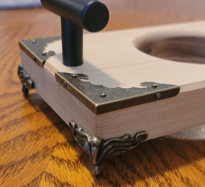 Pu&ograve; includere: Una scatola di legno con una maniglia nera e angoli e piedini in ottone decorati. La scatola ha un ritaglio rotondo nella parte superiore.