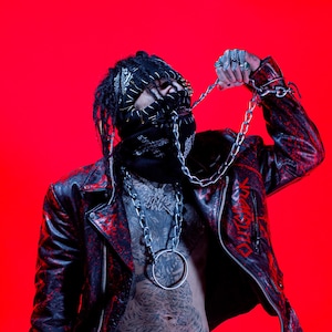 May include: A person wearing a black bandana, a black and red leather jacket, and a silver chain necklace with a large ring. The jacket has red accents and the word "OUTSIDER" in red on the right sleeve. The background is a solid red.